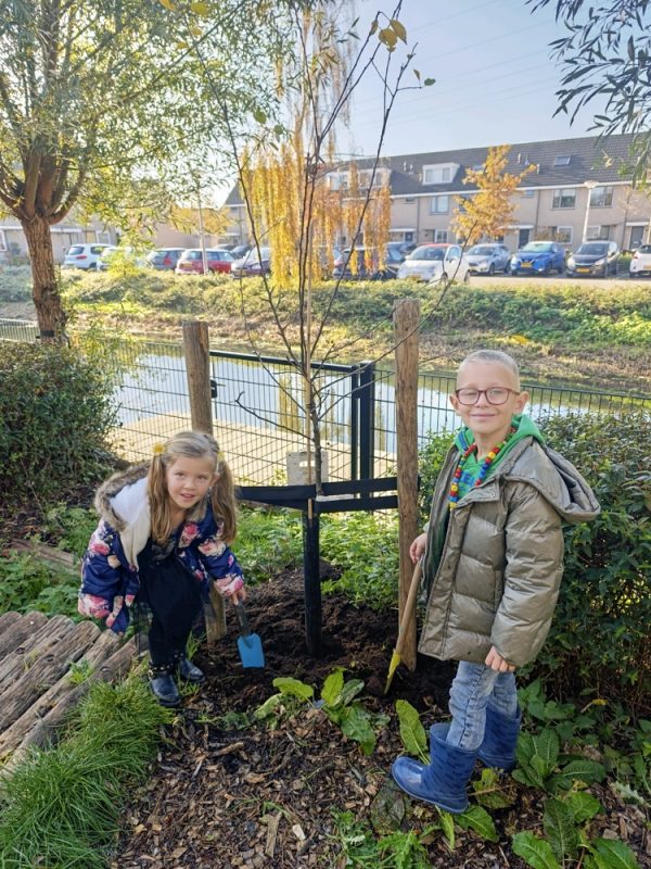 Twee kinderen, een meisje (links) en jongen (rechts) bij een net geplante appelboom in de zijtuin van obs De Tuimelaar.
